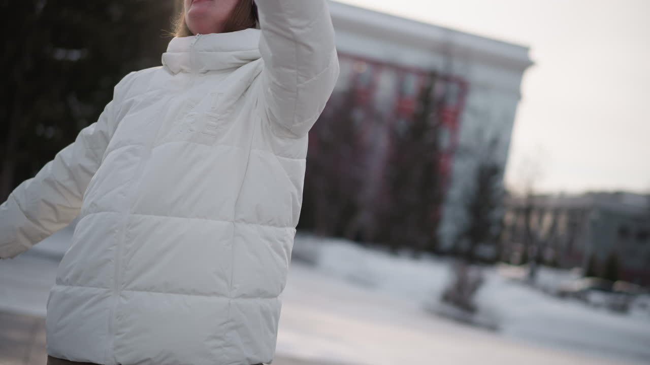 Closeup of youngster wearing white puffer jacket, beanie, black goggles and headphones moving hands and body with rhythmic dance movements on snow covered park plaza under overcast winter light