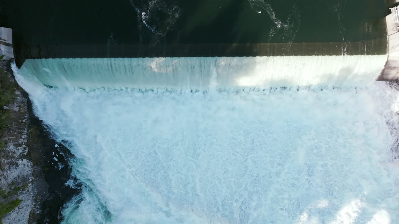 Water spills in a clean line over the top of the dam. The surface above is calm, but below it's all white foam