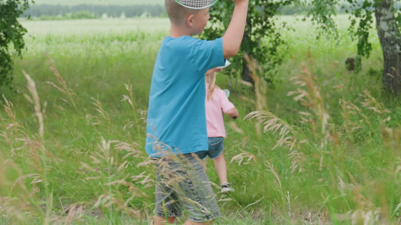 코카서스인 소년과 소녀가 손에 그물을 들고 초원을 달리는 모습, 키 큰 풀과 자작나무 배경, 장난기 넘치는 추격, 웃음과 움직임, 여름 햇살, 솔직한 남매의 유대감, 활기찬 야외 활동, 자연스럽고 편안한 분위기
