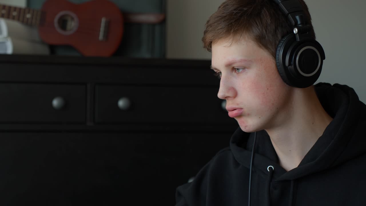 Nærbillede af en ung mand med studiehovedtelefoner, der taler i et musikstudie derhjemme. Kameraet er statisk og fokuseret på hans ansigt med en guitar synlig i baggrunden.