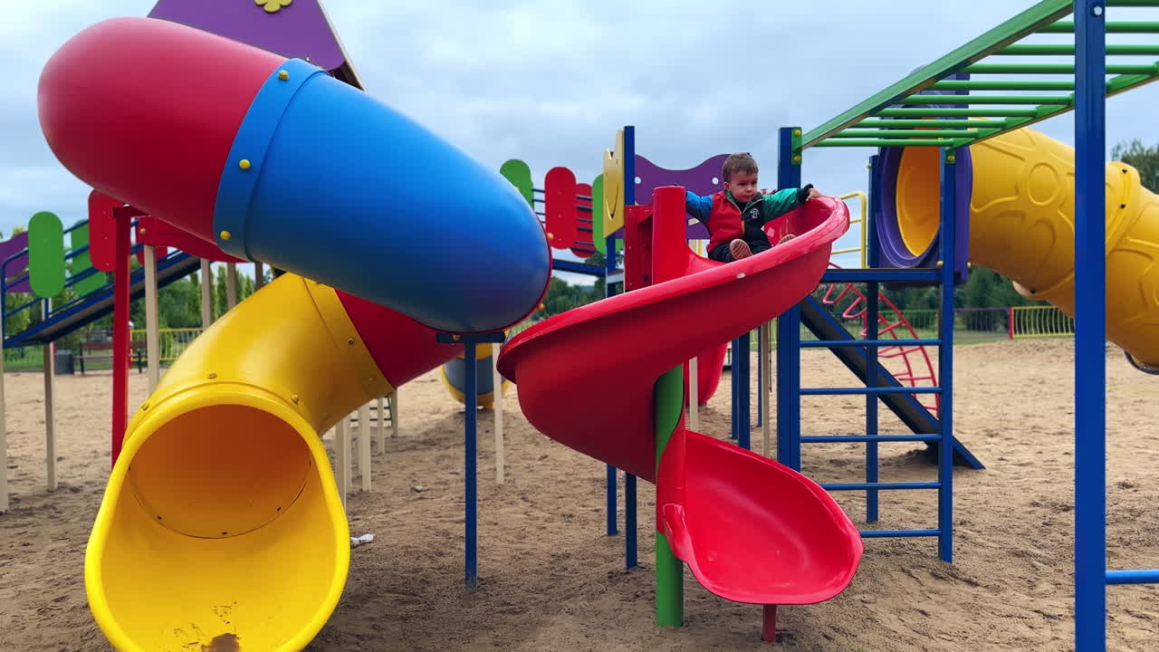 Cute little boy is on the empty playground on grey autumn day. Dark-haired child goes down by the slide.
