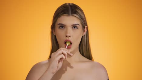 medium static shot of a young pretty woman looking seductively at the camera and licking a vegan lemon-flavored lollipop while enjoying the taste in front of yellow background in slow motion