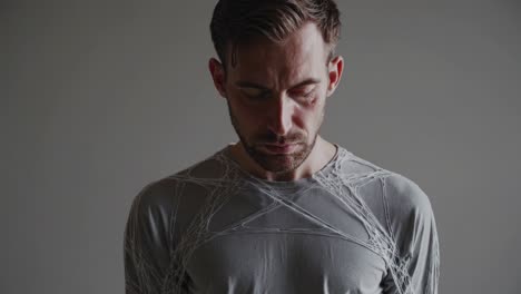 hombre caucásico pensativo con cabello corto y barba marrón, con camisa gris, de pie con la cabeza inclinada y los ojos cerrados contra un fondo neutral, encarnando una profunda introspección