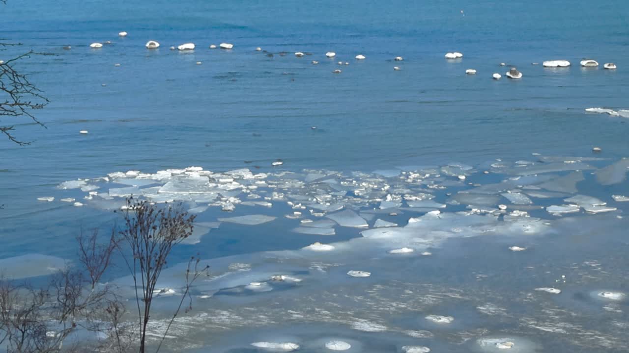 Frozen cold and blue Baltic sea with white ice chunks and pieces in it in Tiskre Estonia during a sunndy day viewed from a high altitude Tabasalu bank cliff though some trees and plants in foreground.