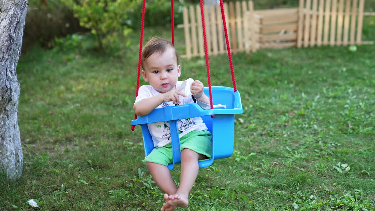 Lovely boy in white t-shirt and green shorts outdoors. Little baby swaying in the plastic swing tied to the tree. Garden backdrop.