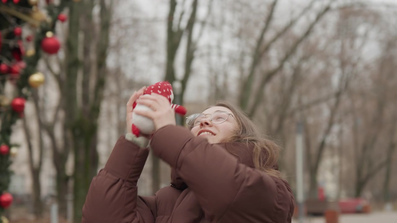 Giovane donna che lancia un nano rosso con un movimento gioioso in un parco innevato, occhiali e piumino marrone visibili tra decorazioni sempreverdi e alberi invernali spogli, sequenza giocosa e spontanea che cattura l'attesa.