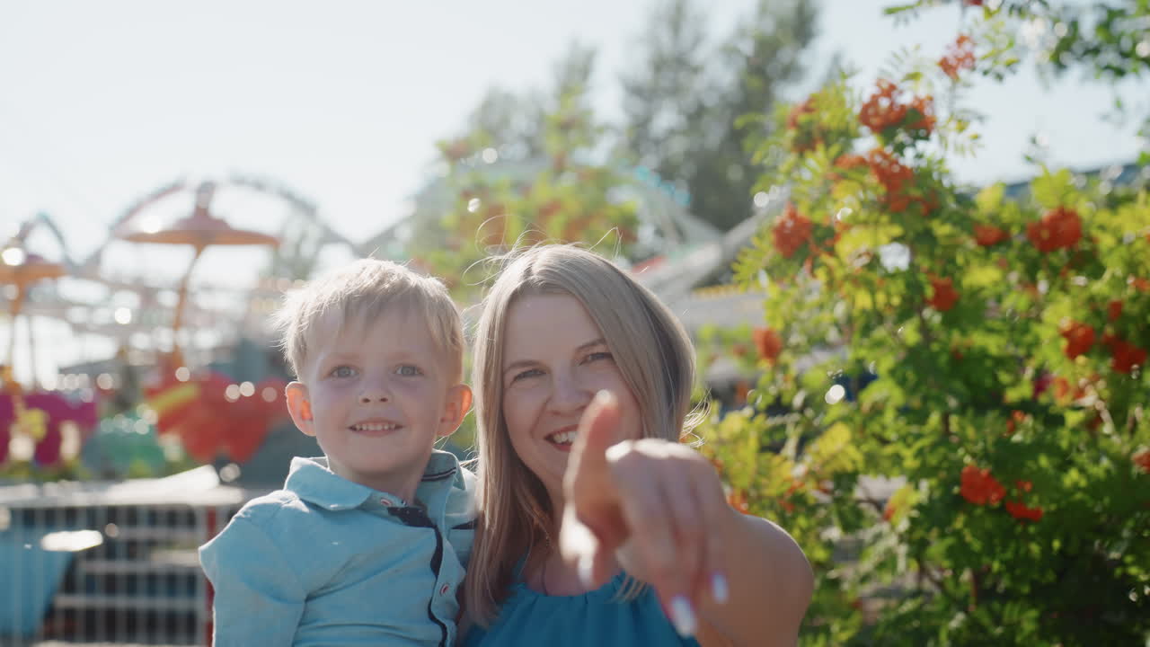 White mother and son in park smiling and pointing at camera among flowers and blurred amusement rides candid weekend family moment with warm sunlight, closeup portrait capturing affectionate bond