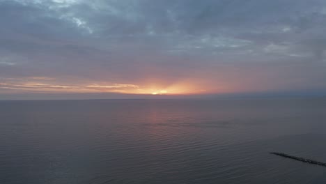 drone-shot-of-calm-sea-during-sunset-with-colorful-clouds-backing-away-revealing-forest-and-shoreline-in-4k