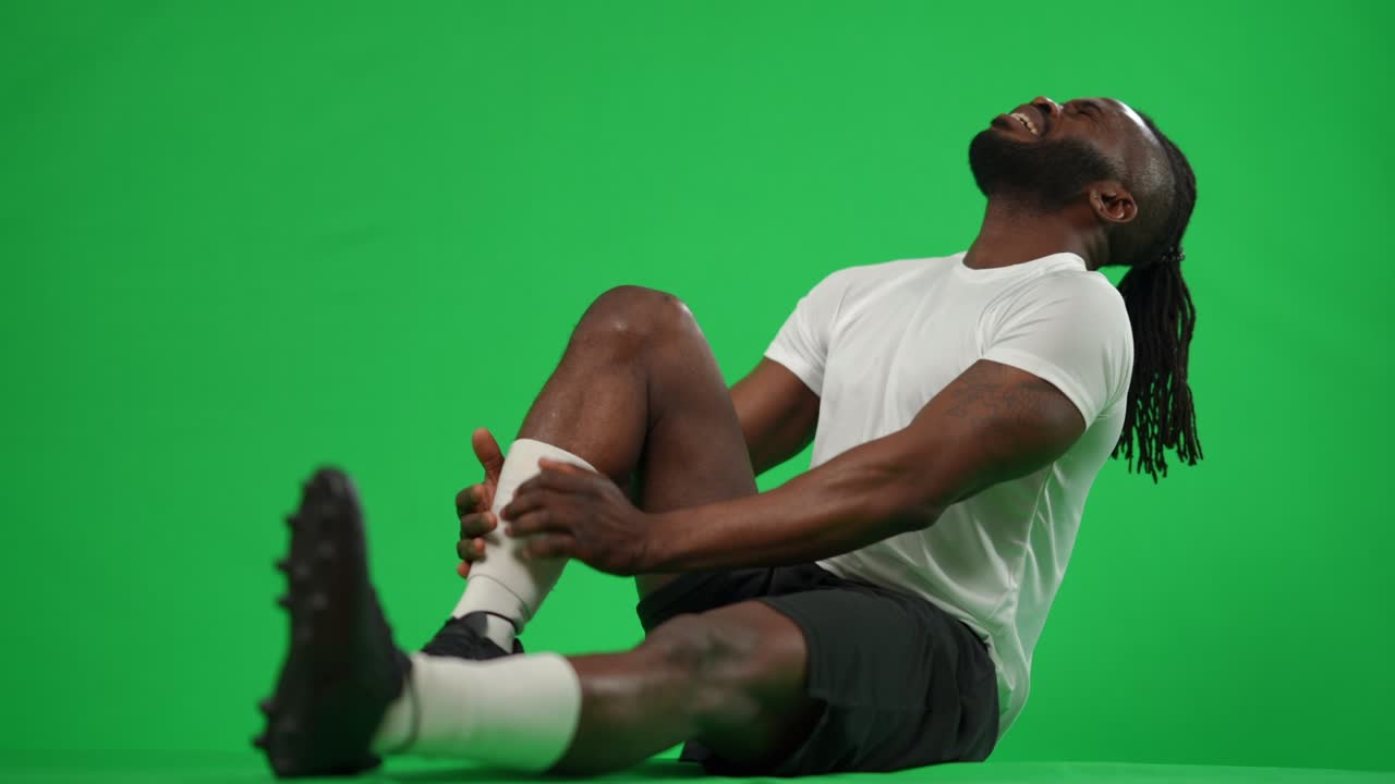 Side view African American footballer sitting on green screen holding painful injured leg. Wide shot portrait of stressed sportsman at chromakey background. Sport trauma concept.