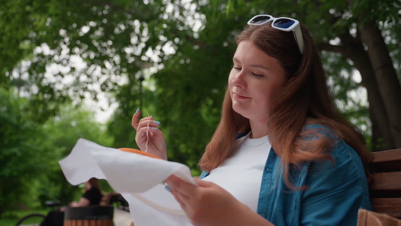 Business woman sitting on wooden park bench focusing on embroidery work with needle and white fabric, wearing white outfit and blue over shirt, enjoying outdoor moment surrounded by fresh green trees
