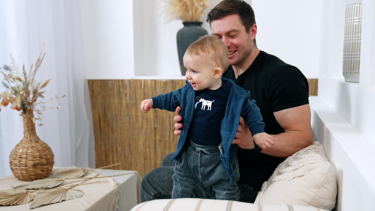 Smiling Caucasian man with baby boy in hands. Little infant learns to stands supported by father.