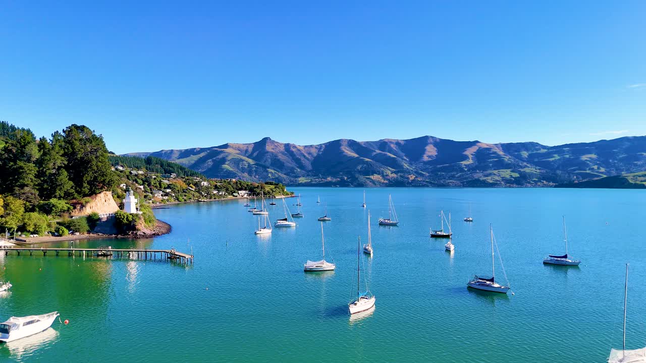 A tranquil scene of Akaroa Harbour with moored boats, lush greenery, and distant hills under clear blue skies