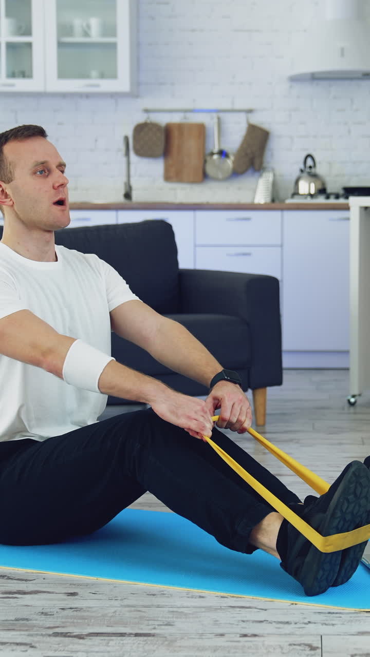 Workout at home during quarantine. Young man is doing breath exercises with fitness rubber band on the floor in front of laptop. Sporty lifestyle indoors. Vertical video