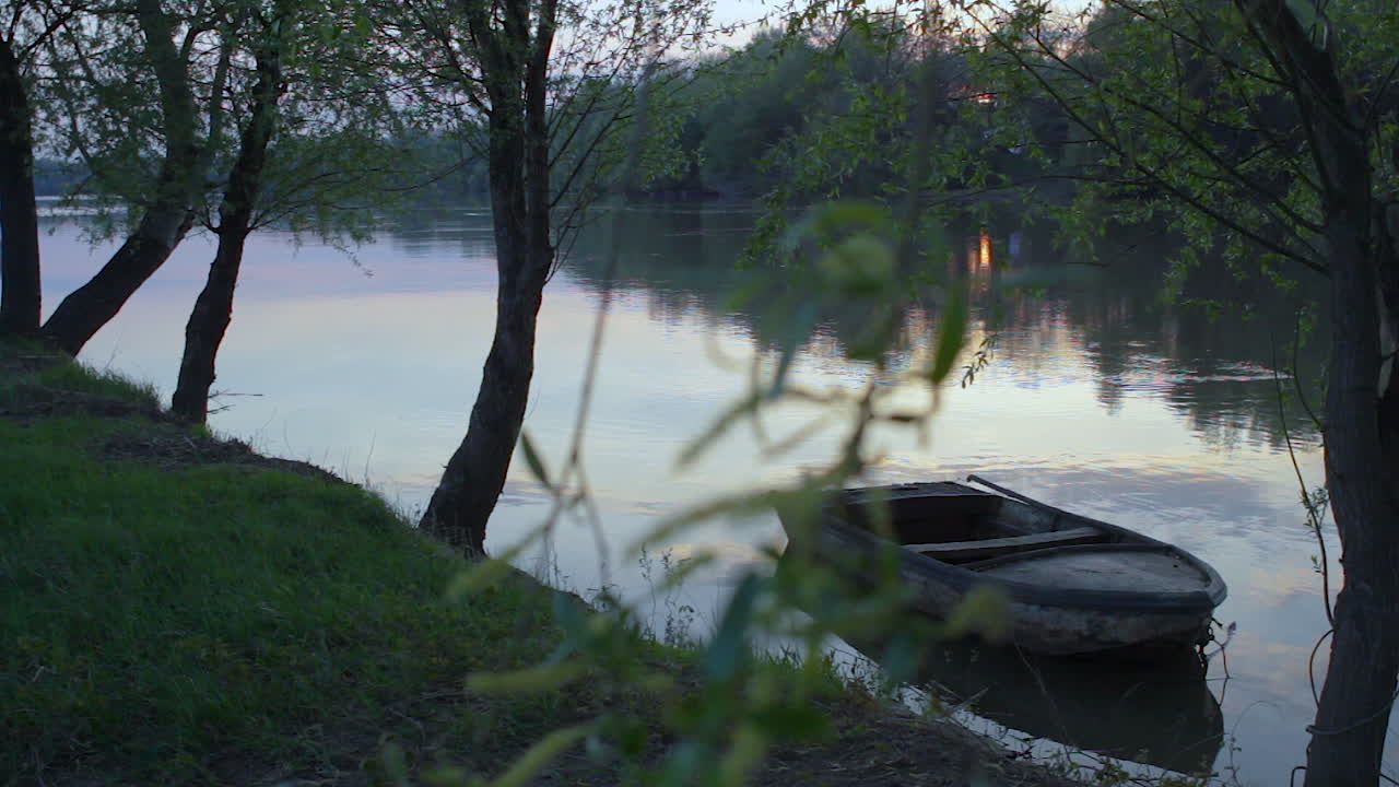 Wide shot of impressionist mood green forest landscape with rusty boat floating on the calmly waving Hungarian Tisza lake in summer time orange sunset