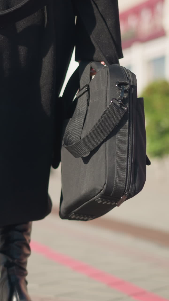 vista trasera de una persona caminando con confianza en una calle urbana iluminada por el sol, con un elegante abrigo negro y botas de cuero, llevando dos bolsas, el fondo borroso muestra el edificio y la planta decorativa