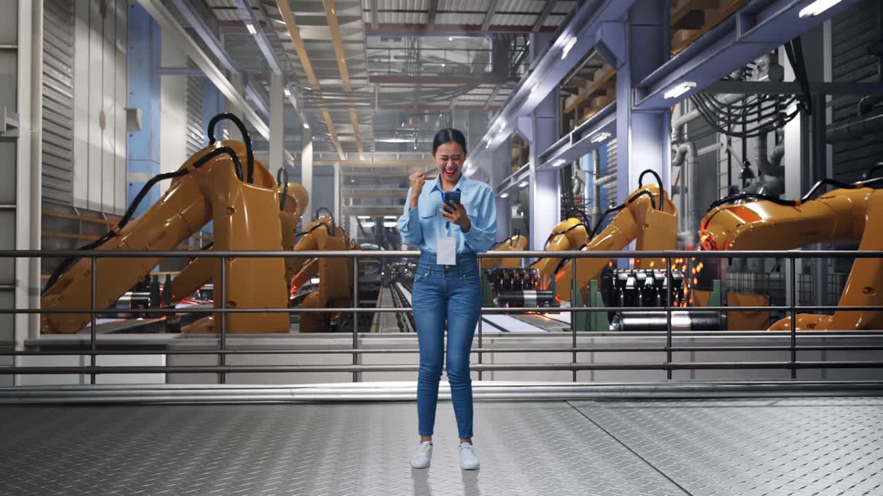 Full Body Of Asian Female With Her Smarphone With Industrial Robotic Arms Working, She Raises Her Fist Up With Screaming Goal