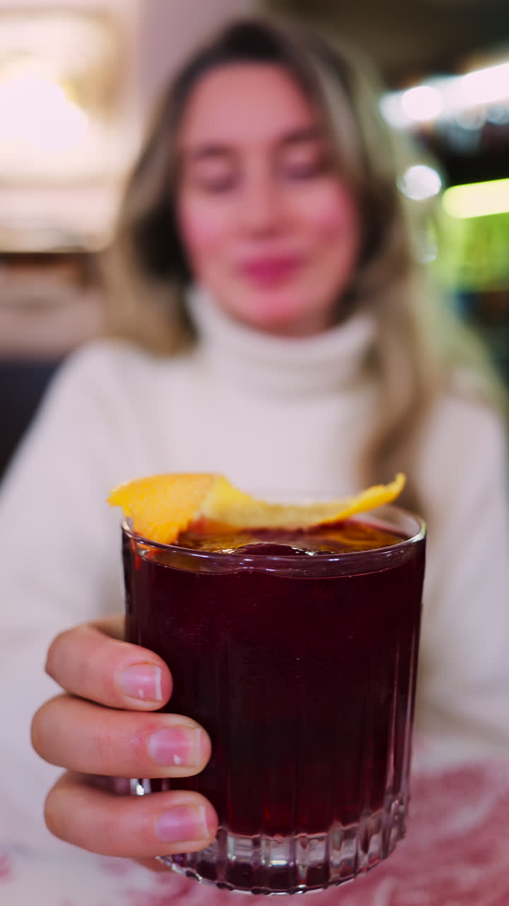 Close up of a woman holding a negroni cocktail on a red and white tablecloth at a restaurant. Vertical