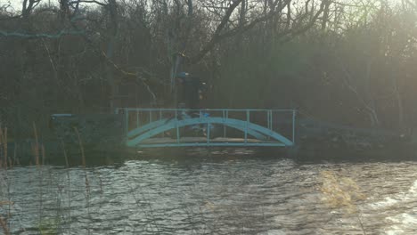 Man-crosses-bridge-cycling-bicycle-along-river-path-in-woods