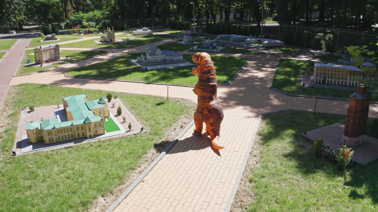 Unknown person in animal suit walking away from camera. Orange dinosaur strolling among mini buildings on beautiful summer day.