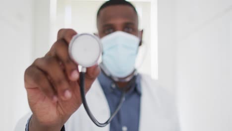 Portrait-of-african-american-doctor-wearing-face-mask-holding-stethoscope-at-home