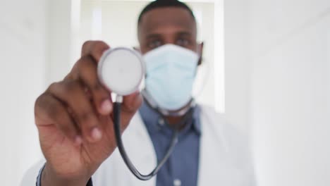 portrait of african american doctor wearing face mask holding stethoscope at home