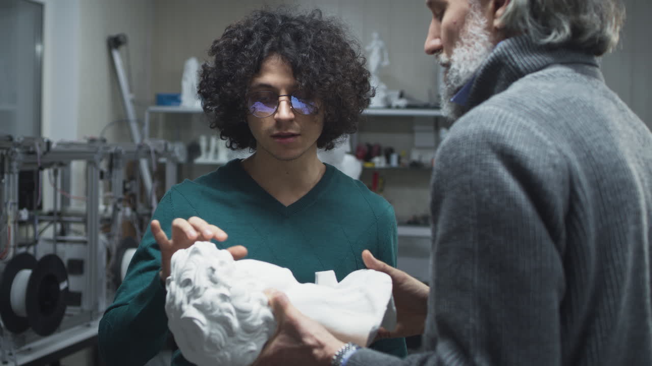 Male Colleagues Discussing Plastic Bust. Young and mature men in casual clothes examining and discussing plastic bust while working in 3D printing studio together