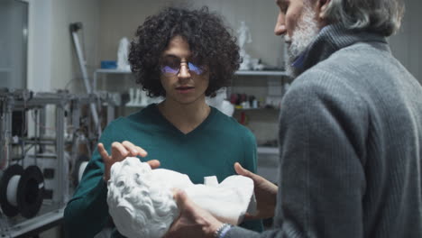 male colleagues discussing plastic bust. young and mature men in casual clothes examining and discussing plastic bust while working in 3d printing studio together