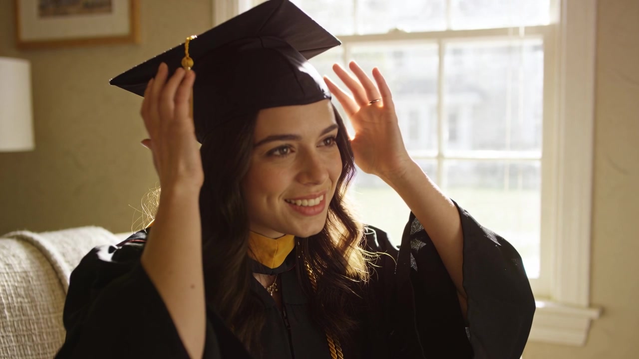 Donna adulta che prende e sistema il tocco sul divano di casa per la laurea, indossando la toga e una nappa dorata