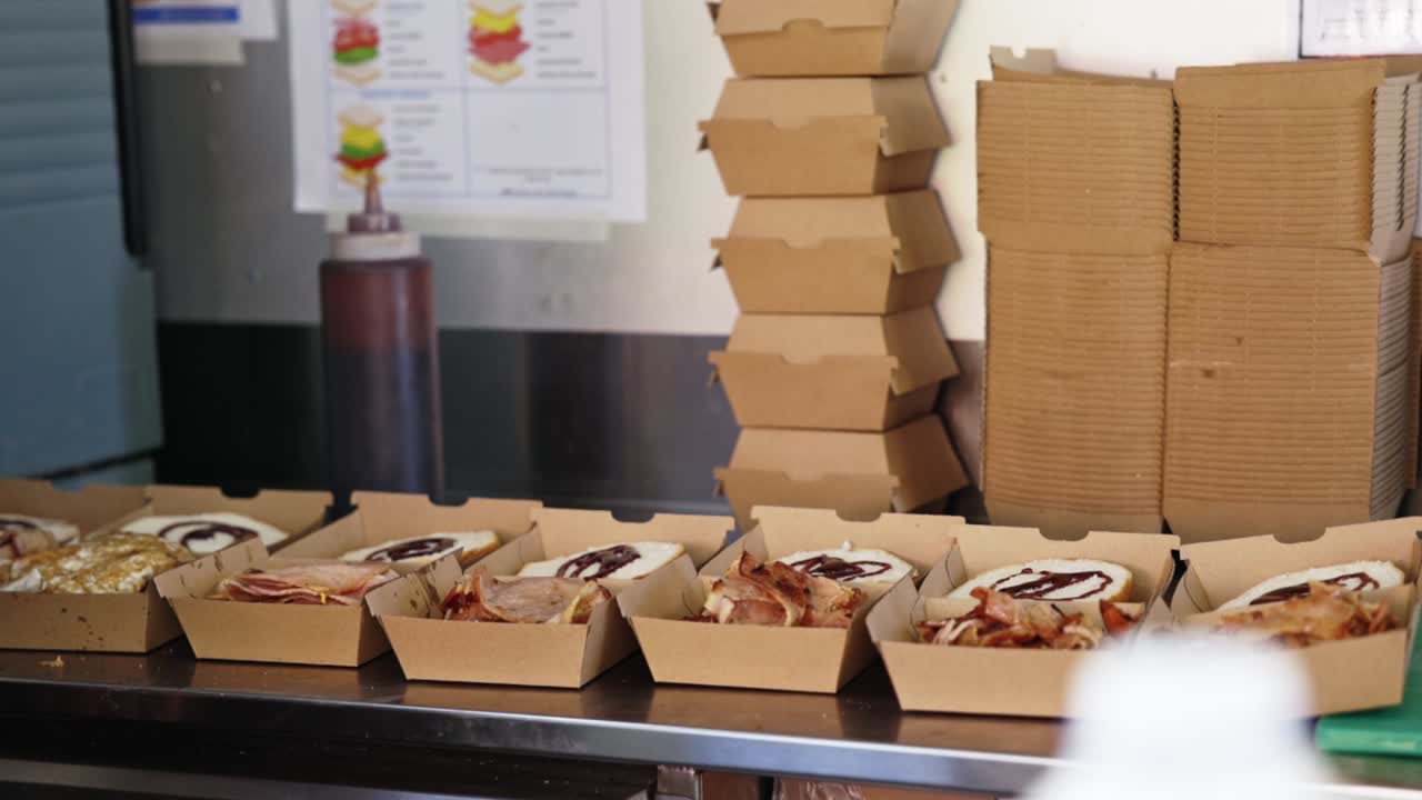 Close-up de um funcionário de food truck montando rolos de bacon e ovo em caixas para viagem, preparando pedidos em uma cozinha móvel movimentada.