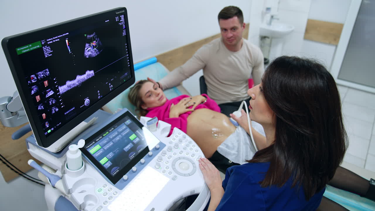 Female doctor sits in front of ultrasound machine. Medic scans pregnant lady on the couch. Caucasian man strokes his wife's head supportively. Top view.