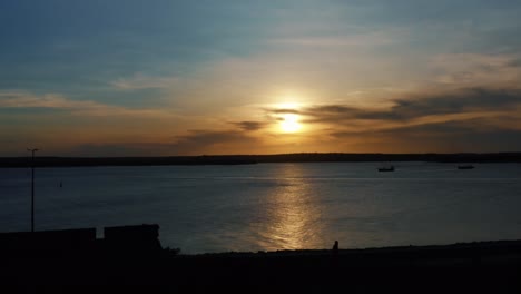 Stunning-aerial-drone-dolly-out-shot-of-a-golden-sunset-reflecting-onto-the-water-and-the-camera-passing-over-a-famous-historical-military-fort-in-Joao-Pessoa,-Brazil