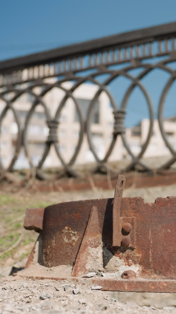 Close view of a rusty metal object lying on the ground with an ornate urban fence in the background, with cars passing on the road