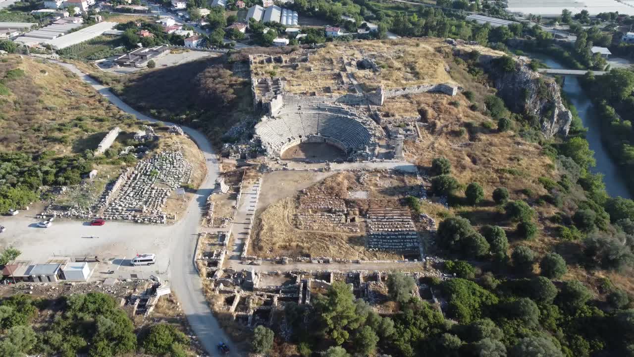 Aerial drone view showing semi circular amphitheater with surrounding stone ruins of ancient city Myra in Antalya Province under bright daylight across dry terrain mixed with patches of greenery