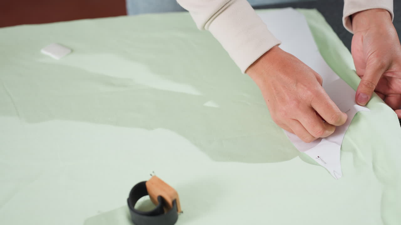 creative hands pinning paper design pattern onto light green fabric on workspace table while magnetic wrist tool for pins lies nearby, showcasing fashion preparation process in bright sewing studio