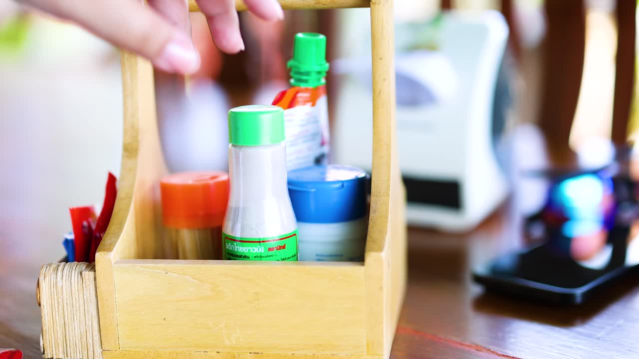 una mano alcanza una botella de condimentos de un caddy de madera en una mesa en un restaurante de phuket. la iluminación brillante realza la escena