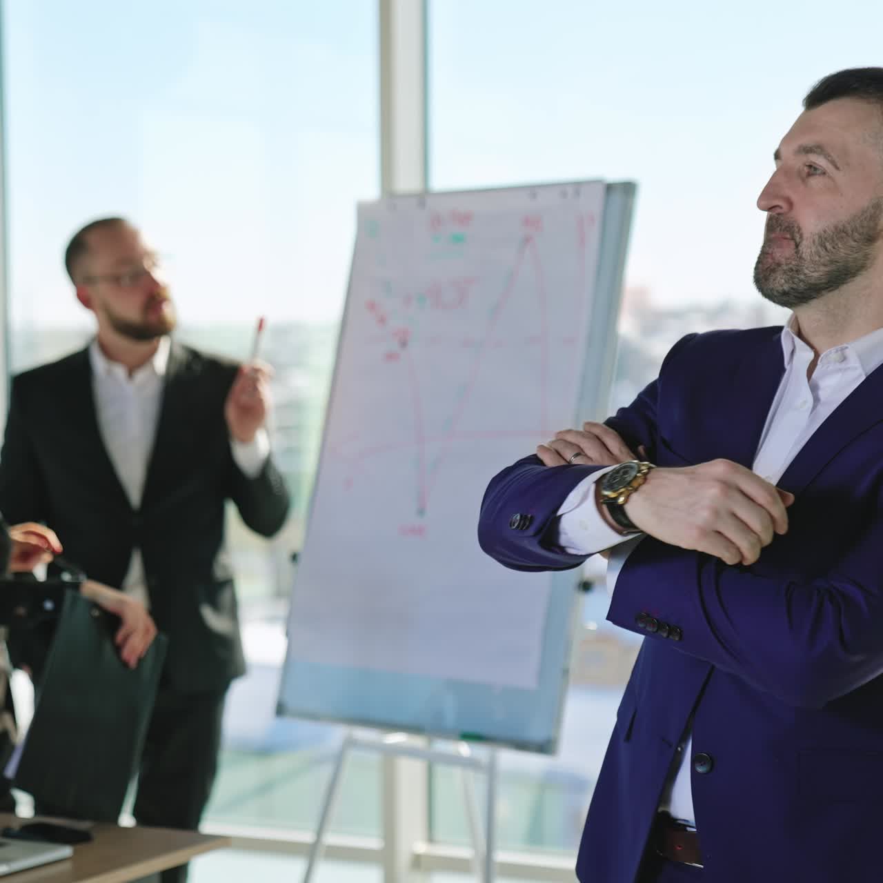Mid-aged bearded man in suit talks firstly to camera and then aside. Portrait of a confident male business person crossing hands on his chest. Office employees at backdrop in blur
