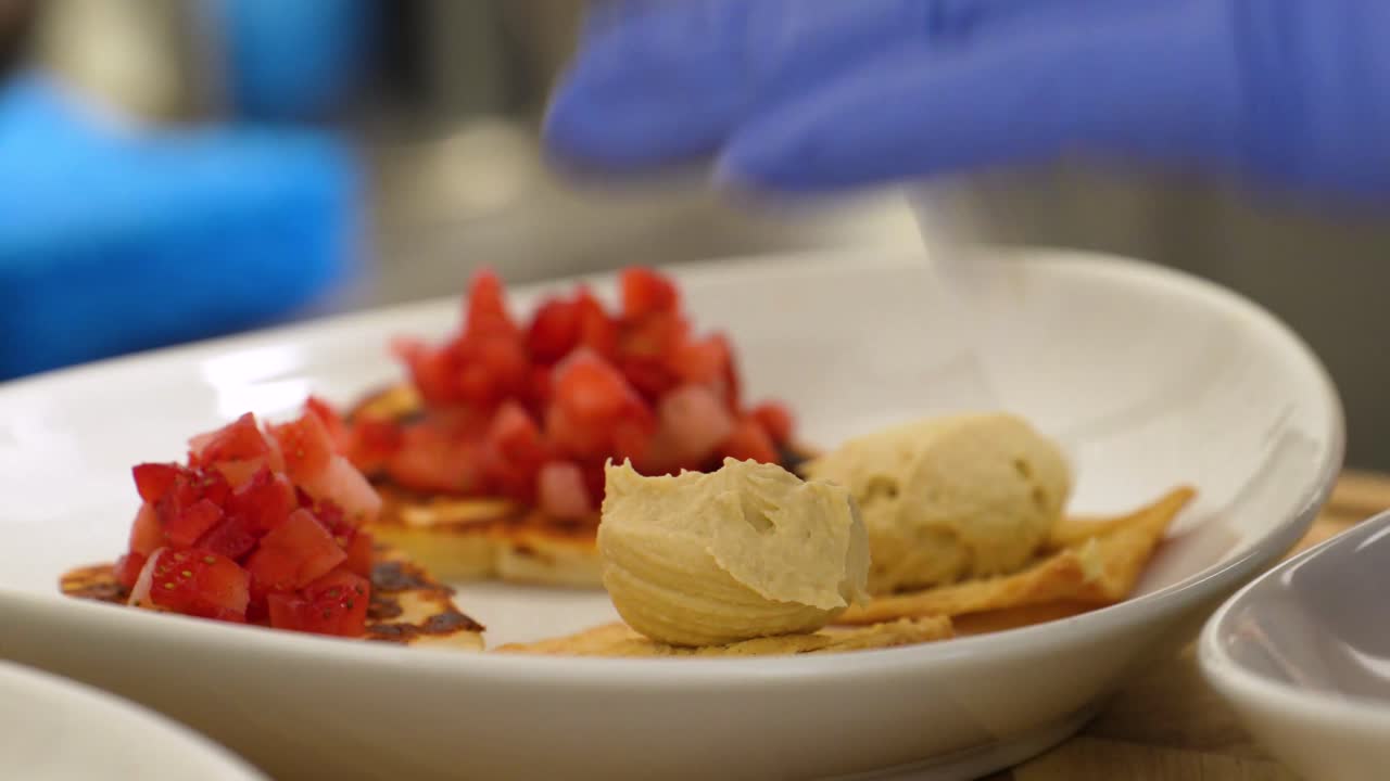 Cook Wearing Blue Rubber Gloves Places Hummus on Pita Bread Chips with Diced Tomatoes, Assembling a Healthy Appetizer Meal by in the Kitchen using a Spoon and White Bowls or Plates, Food Closeup