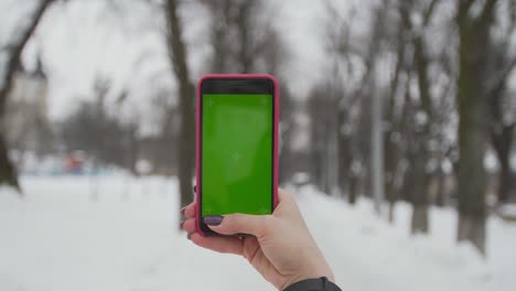 young woman types and slides on her smartphone in the winter park. cellphone with chroma key screen - green screen.close up. tracking motion.