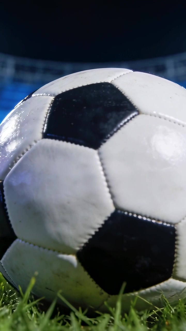 Dikey video: Suni çimdeki futbol topuna doğru yakınlaşan hareketli kamera, projektör yansımalarını ortaya çıkarıyor.