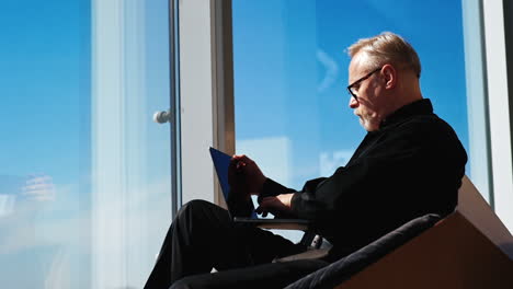 focused male wearing black shirt and trousers sits at the window. man works at laptop typing on keyboard and closing the gadget.
