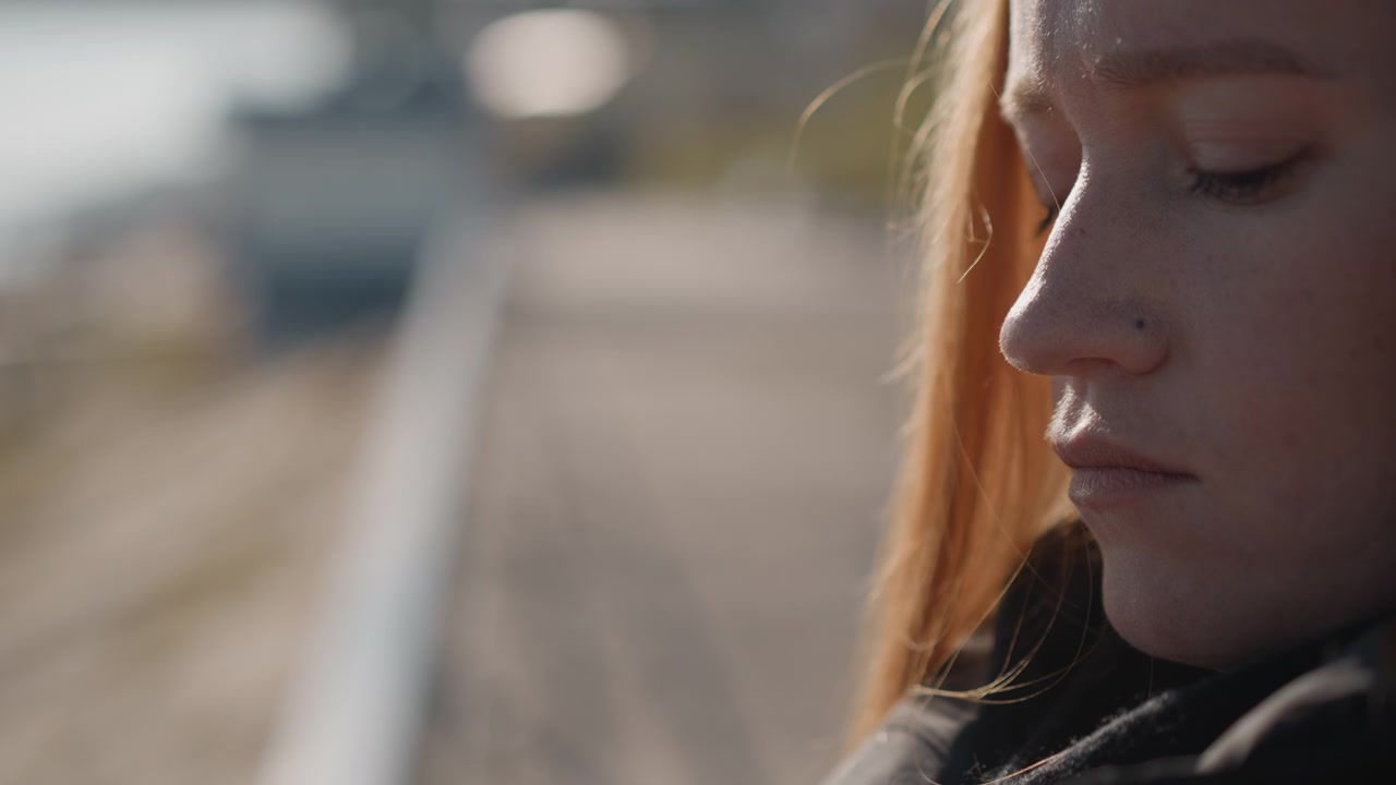 Una donna con i capelli lunghi si sistema la giacca nera all'aperto, concentrandosi sul movimento delle mani e del tessuto. La luce naturale soffusa evidenzia i dettagli della texture contro uno sfondo sfocato con elementi distanti.