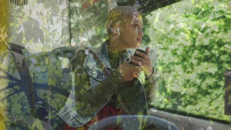 Animation-of-leaves-over-biracial-woman-sitting-in-bus,-using-smartphone