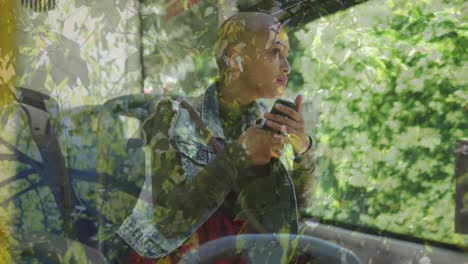 animation of leaves over biracial woman sitting in bus, using smartphone