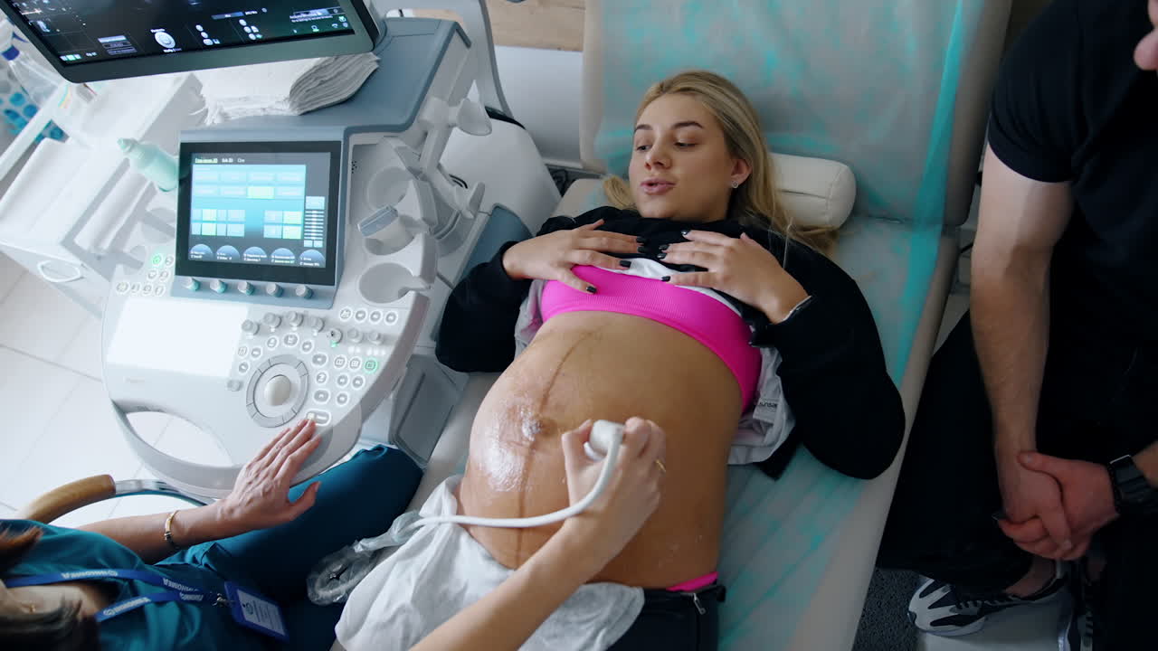 Pregnant Caucasian lady with big belly has ultrasound diagnostic. Doctor moves device by the gelled abdomen. Top view.