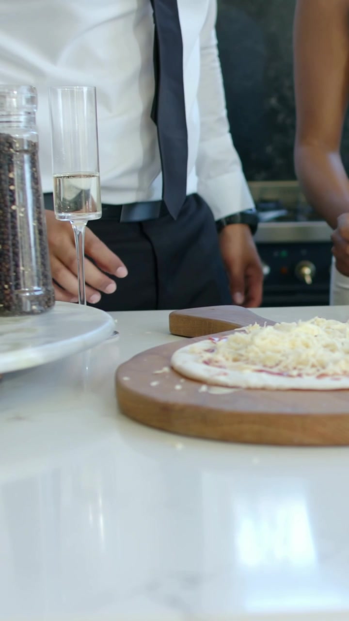 Vertical video: Woman in tan dress reaching pizza board, man placing flute on counter serving pizza