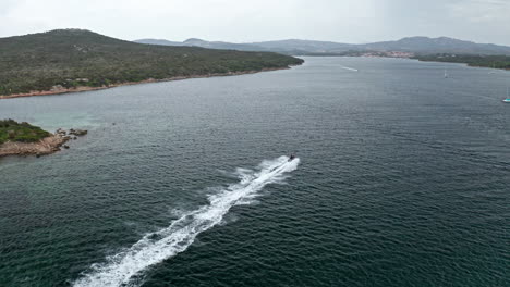 A-speedboat-cruises-across-clear-blue-waters-near-the-lush,-green-coastline-of-Sardinia