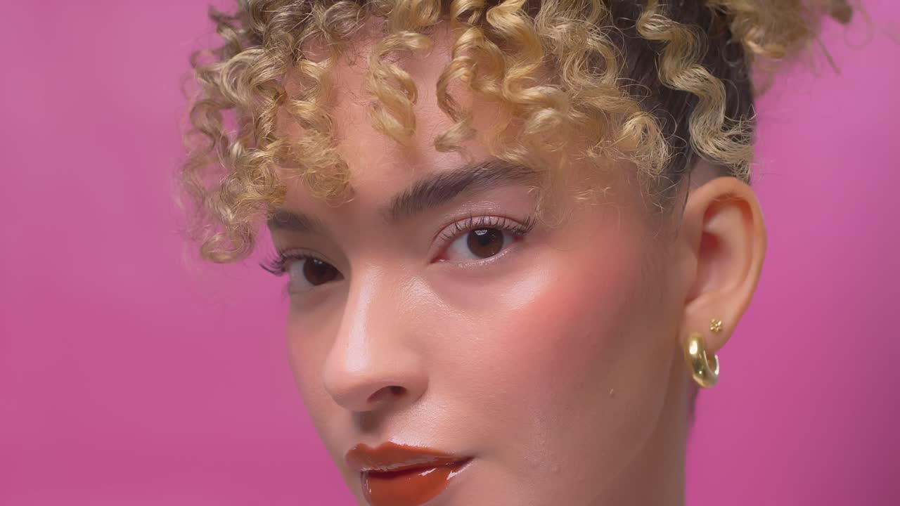 Close up of young woman with curly hair posing against a pink backdrop under soft lighting