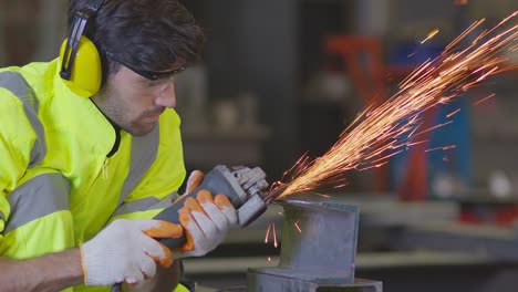 trabajador masculino caucásico en un uniforme seguro usar soldadores cueros, rueda eléctrica molienda en la estructura de acero en el factor en el garaje, chispas bokeh naranja vuelan a los lados