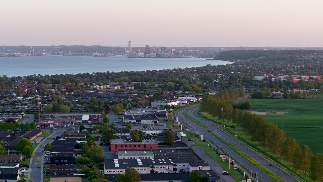 Aerial drone footage overlooking a suburban neighborhood with a main road, trees lining the avenue, and a coastal city skyline in the distance under early morning light