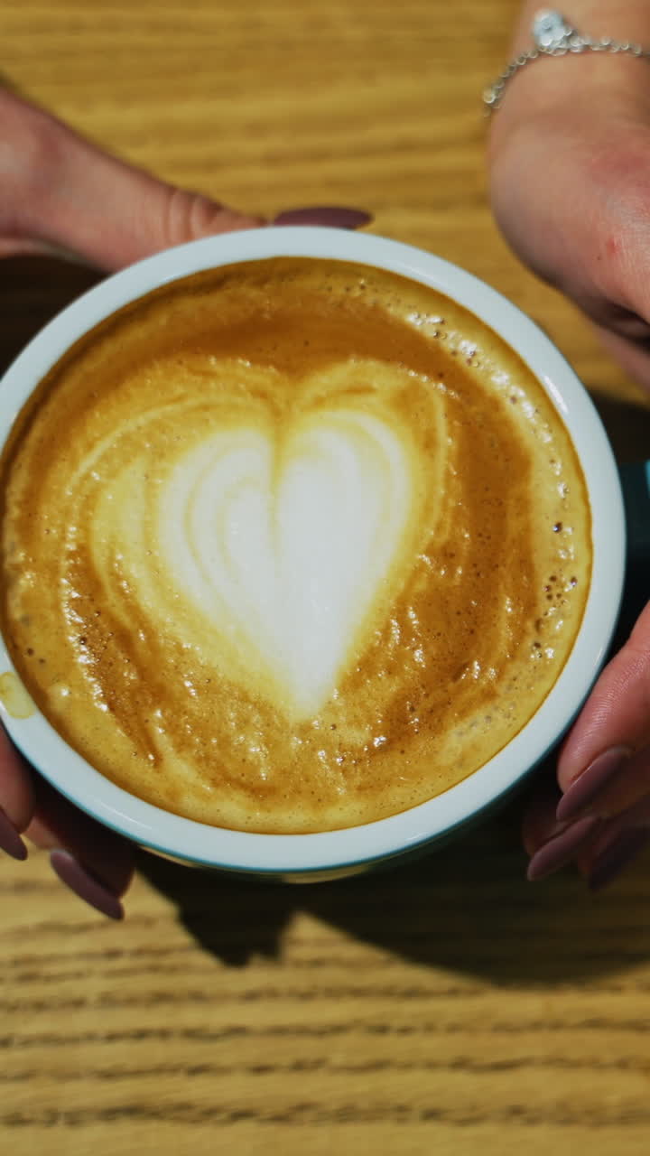 Cup with coffee on wooden background. Latte art of heart design on coffee. Female hands taking cup of hot drink from the wooden surface. Top view. Vertical video